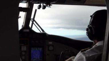 Pilotlar deniz uçağının kontrolünde oturuyorlar. Pencerenin manzarası gri bulutlar ve turkuaz okyanus sularıdır. Paneldeki farklı düğmeler yanıyor. İki pilot uçağı kontrol ediyor.