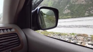 Dikiz aynası ve dağ nehri manzarası. Aynaya beyaz bir SUV yansıtılır, ve uzakta kayalık kıyıları olan bir nehir görebilirsiniz. Geçitten geçen off-road. Tehlikeli bir macera.