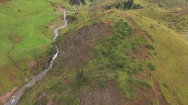 FPV İHA 'sını nehir kıyısında uçurmak. Yeşil tarlalar ve dağlar, çalılar ve kozalaklı ağaçlar her yerde yetişir. Küçük bir şelalesi olan kaynayan bir nehir. Bulutlu, bulutlu ve hafif yağmurlu.