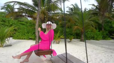 Pembe beyaz şapkalı bir kız sahilde salıncakta sallanıyor. Beyaz mercan kumu, bulutlu karanlık gökyüzü. Kız elinde bir kokteyl tutuyor. Turkuaz okyanus suyu ve yeşil palmiye ağaçları. Rahatlama