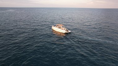 Sersemletici bir günbatımında sakin, koyu mavi bir denizde süzülen küçük beyaz motorlu tekne, gökyüzü turuncu ve pembe renklerle dönüşürken ardında hafif bir uyanış bırakıyor.