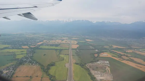 Tarımsal bir arazinin üzerinde uçan bir uçak kanadı tarlaların, dolambaçlı yolların üzerinde süzülüyor ve arka planda berrak bir gökyüzünün altında yükselen görkemli dağlarla dolu tuhaf bir köy.