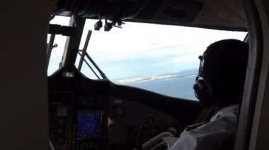 Pilotlar deniz uçağının kontrolünde oturuyorlar. Pencerenin manzarası gri bulutlar ve turkuaz okyanus sularıdır. Evler ve bir ada uzaktan görülebilir. Uçak iniyor.