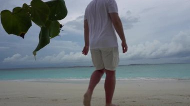 Beyazlı bir adam sahilde yürüyor ve heyecanlanıyor. Turkuaz berrak su, gökyüzünde kara bulutlar ve beyaz mercan kumu. Ağaçtan yapraklar. Kumda ayak izleri bırakıyor. Hafif meltem ve yağmur