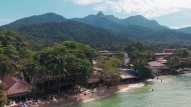 Turkuaz okyanus suyu, turistlerin rahatlayıp tropikal manzaranın tadını çıkardıkları Koh Chang adasının kumlu sahillerinde yavaşça uzanır. Arka planda yemyeşil dağlar görkemli bir şekilde yükselir.