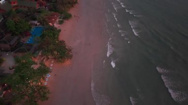 Tropikal koh chang adası plajı gün ışığından akşama geçiş yapıyor, ıslak kumda yürüyen turistleri gözler önüne sererken, sakin günbatımı anında hafif dalgalar kıyıları okşuyor.