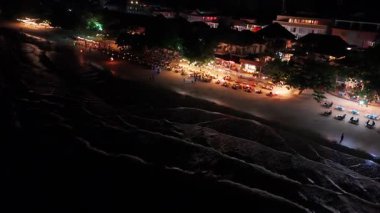 Aydınlatılmış restoranlar ve barlarla dolu canlı bir sahil gece hayatı, okyanus suları yakınında dinlenen turistler, eğlence ve eğlence dolu bir akşam ortamı yaratıyor.