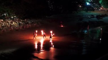 Geceleri Tayland 'da bir plajda ateş dansı yapan turistler, kumların üzerine kurulmuş masa ve sandalyelerle canlı ve şenlikli bir atmosfer yaratıyorlar.