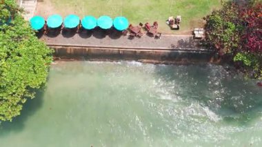 Turkuaz okyanus, Koh Chang adasının kumlu kıyılarında, turistler mavi şemsiyeler altında dinlenirken ve arka planda görkemli bir şekilde yemyeşil dağlar yükselirken.