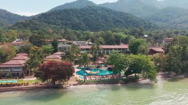 Yüzme havuzu ve plaj sandalyeleri olan Koh Chang Adası tatil köyü bazı kırmızı ağaçların altında, ormanlı dağlar ve arka planda turkuaz su ile