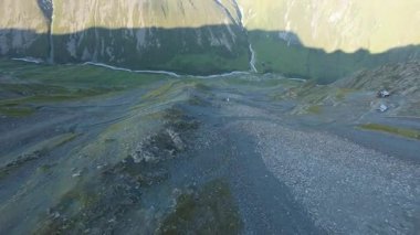 Vadideki dik uçurumlar boyunca bir FPV insansız hava aracına atlamak. Güneş ışınları tepenin arkasından geliyor. Büyük kayalar, yeşil çimenler ve vadiden aşağı akan bir nehir. Dağların arasında uçarak