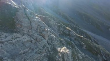 Vadideki dik uçurumlar boyunca bir FPV insansız hava aracına atlamak. Güneş ışınları tepenin arkasından geliyor. Büyük kayalar, yeşil çimenler ve vadiden aşağı akan bir nehir. Dağların arasında uçarak