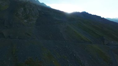 Vadideki dik uçurumlar boyunca bir FPV insansız hava aracına atlamak. Güneş ışınları tepenin arkasından geliyor. Büyük kayalar, yeşil çimenler ve vadiden aşağı akan bir nehir. Dağların arasında uçarak