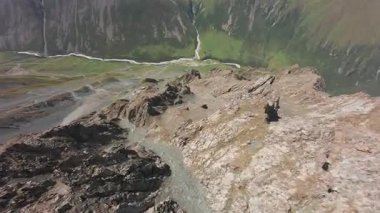 Vadideki dik uçurumlar boyunca bir FPV insansız hava aracına atlamak. Güneş ışınları tepenin arkasından geliyor. Büyük kayalar, yeşil çimenler ve vadiden aşağı akan bir nehir. Dağların arasında uçarak
