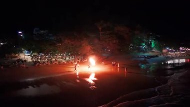 Gece sahnesinde canlı bir yangın performansı yakalayan turistler, renkli restoranlar ve bar aydınlatması arasında alevlerle hareket eden dansçıları seyreden turistler.