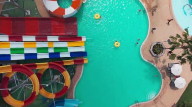 Canlı bir su parkının yüksek açılı görüntüsü, turkuaz havuzlarda yüzen, kayan ve dinlenen turistleri renkli slaytlar ve çeşitli gösteriler arasında gösteriyor.