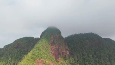 Verdant dağ yamaçları yoğun tropikal ormanlarla örtülmüş, bulutlu gökyüzünün altında uzanan katmanlı yeşil tepeleri ortaya çıkarıyor, el değmemiş vahşi doğayı gözler önüne seriyor.