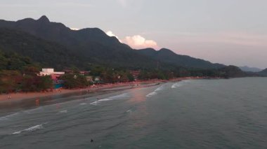 Koh Chang adası plajı güneşin sıcacık parıltısıyla yıkanır, turistler kumsalda dinlenir ve yemyeşil dağların çerçevelediği sakin denizde yüzerler.