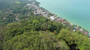 Bereketli bitki örtüsü yamaçları kaplıyor canlı bir kıyı şeridi ile buluşuyor, binalar ve yollar yeşillikler arasında görülebiliyor, dinamik bir fpv uçuş manzarası sunuyor