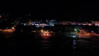 Gece sahili şenliklerinde enerjik bir kalabalık renkli aydınlatma altında dans ediyor, okyanus kıyısında elektrik atmosferi yaratıyor ve kutlamaları gerçekleştiriyor.