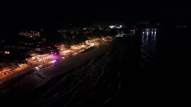 Sıcak ışıklar, hareketli bir sahil beldesini gece aydınlatıyor. Turistler kumlu sahilde geziniyor ve okyanusun nazik dalgaları büyüleyici bir atmosfer yaratıyor.