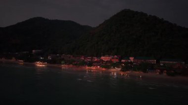 Sıcak parti ışıkları, tropikal kıyı şeridi boyunca gece vakti okyanus sularını aydınlatarak, akşam alacakaranlığı sırasında dingin koh chang adası manzarasında canlı yansımalar oluşturur.