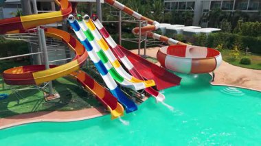 Yüzme havuzunun yanında kıvrılan parlak, renkli su kayakları yeşil manzaralı ve mavi gökyüzü olan boş su parkında canlı su canlılığı sergiliyor.