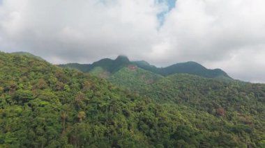 Verdant dağ yamaçları yoğun tropikal ormanlarla örtülmüş, bulutlu gökyüzünün altında uzanan katmanlı yeşil tepeleri ortaya çıkarıyor, el değmemiş vahşi doğayı gözler önüne seriyor.