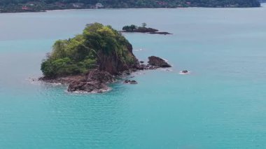 Kristal berrak turkuaz su, bereketli bitki örtüsüyle kaplı küçük, kayalık bir adanın çevresini sarar.