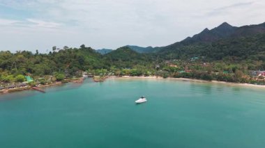 Turkuaz su, yumuşak bir gökyüzü altında yemyeşil ve uzak dağların çerçevelediği sakin manzaraya lüks bir dokunuş katan beyaz bir yat, tropikal bir adanın kumlu sahilini nazikçe kaplar.