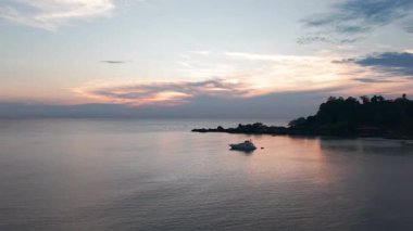 Sakin okyanus suyu günbatımının pembe, turuncu ve mor renklerini yansıtır. Beyaz bir yat, tropik bir ada kıyısında huzur içinde süzülür. Huzurlu ve pitoresk bir manzara yaratır.