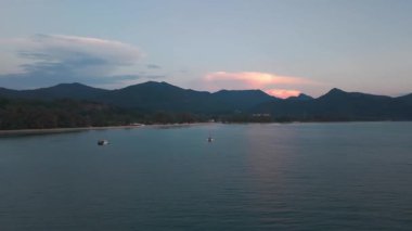 Canlı pembe ve turuncu bulutlar uzak dağ siluetleri üzerinde sürüklenirken sakin tropikal körfez sularında yüzen balıkçı tekneleri akşam alacakaranlığı sırasında