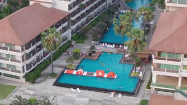 Palmiye ağaçları ve otel binalarıyla çevrili tropikal bir tatil köyünde yüzen ve havuzda oynayan turistlerin manzarası arttı.