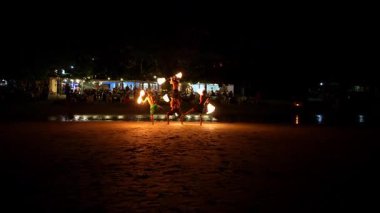 Çevik ateş dansçıları yeteneklerini kumlu bir plajda sergiliyorlar, sakin bir gecenin arka planında yanan meşalelerinin sıcacık parıltısıyla aydınlanıyorlar.