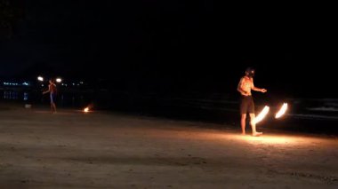 Yetenekli ateş hokkabazları yeteneklerini kumlu bir plajda sergiliyor. Gecenin karanlığına karşı meşalelerinin büyüleyici alevleriyle aydınlanıyor.