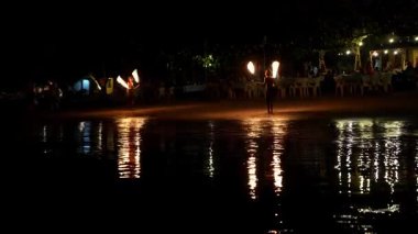 Profesyonel ateş dansçısı büyüleyici bir gece feneri hokkabazlığı yapıyor dinamik alevler saçıyor ve koyu kumlu sahil şeridinde parıldayan yansımalar yapıyor.