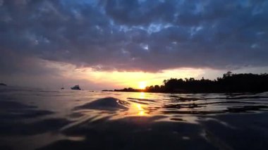Altın saat güneş ışığı, tropikal okyanusun nazik dalgalarını yansıtır. Uzak teknelerle ve dramatik bulutlu gökyüzüyle pitoresk bir sahne yaratır.
