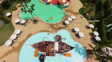 Renkli su kaydırakları, korsan gemisi oyun parkı, güneş pansiyonları ve şemsiyeleri olan bir tatil köyünde oynayan ailelerin manzarası yükseldi.