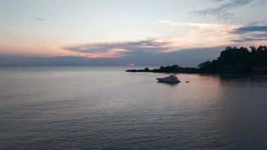 Sakin okyanus, güzel günbatımı renklerini yansıtıyor. Tropikal bir adanın kıyısında yüzen bir yat huzurlu ve güzel bir manzara yaratıyor.