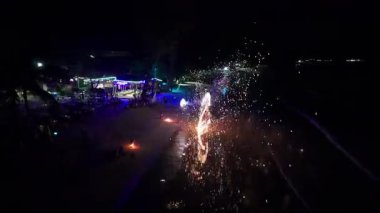 Gece sahnesinde yetenekli ateş dansçıları tropik tatil köyünün yakınında büyüleyici bir dönüş gösterisi yapıyor, turistleri göz kamaştırıcı alevlerle eğlendiriyor ve karanlık okyanus zeminine karşı kıvılcımlar saçıyor.