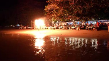 Ateş dansçısı, deniz kenarı restoranı yakınlarında büyüleyici bir gece gösterisi sergiliyor. Turistleri eğlendirirken ıslak kumu yansıtıyor. Tropikal akşam gökyüzü altında içkilerinin keyfini çıkarıyor.