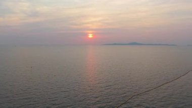 Ilık güneş ışığı sakin okyanus yüzeyine yansıyor. Batan güneşe doğru parıldayan bir yol yaratıyor. Su boyunca uzanan bir şamandıra hattı ve ufukta görünen uzak bir adayla.