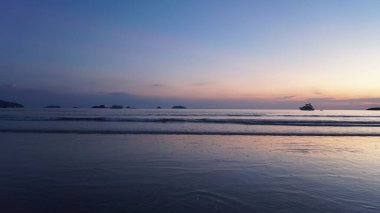 Gün batımında kıyıya vuran sakin deniz ufukta kayıklar ve adalar, sakin ve huzurlu bir atmosfer yaratıyor.