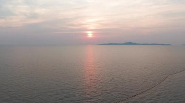 Sakin okyanus suları canlı turuncu günbatımı ışığını yansıtıyor, uzak ada silueti, sakin su yüzeyinde uzanan şamandıra çizgisi ile altın ufuk çizgisine karşı.