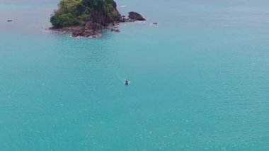 Kristal berrak turkuaz su, bereketli bitki örtüsüyle kaplı küçük, kayalık bir adanın çevresini sarar.