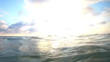 Güneş ışığı bulutları ve okyanus dalgalarını aydınlatır, yavaş yavaş su altı perspektiflerine geçiş yapar, ışık yansımalarını ve suların derinliklerini yakalar.