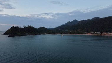 Sakin okyanus suları, güneş ışığının sıcak tonlarını yansıtıyor. Görkemli dağlar ve el değmemiş bir plaj tarafından çerçevelenmiş. Huzurlu ve sakin bir atmosfer yaratıyor.