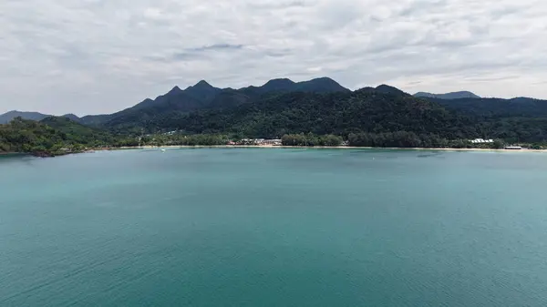Turkuaz su, tropikal bir adanın kumlu sahilinde yavaşça dolaşır, yemyeşil dağlar bulutlu bir gökyüzünün altında ihtişamlı bir şekilde yükselir, sakin ve resimsel bir manzara yaratır.
