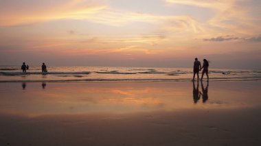 Gün batımında ıslak bir plajda yürürken el ele tutuşan çift. Arka planda yıkanan turistler ve sakin su yüzeyine yansıyan titrek gökyüzü.