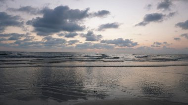 Yumuşak dalgalar yavaşça kumlu kıyıya vuruyor güneş ufkun altına batarken, plajda sakin bir günbatımında, turuncu ve pembe renkli canlı gökyüzünü boyuyor.